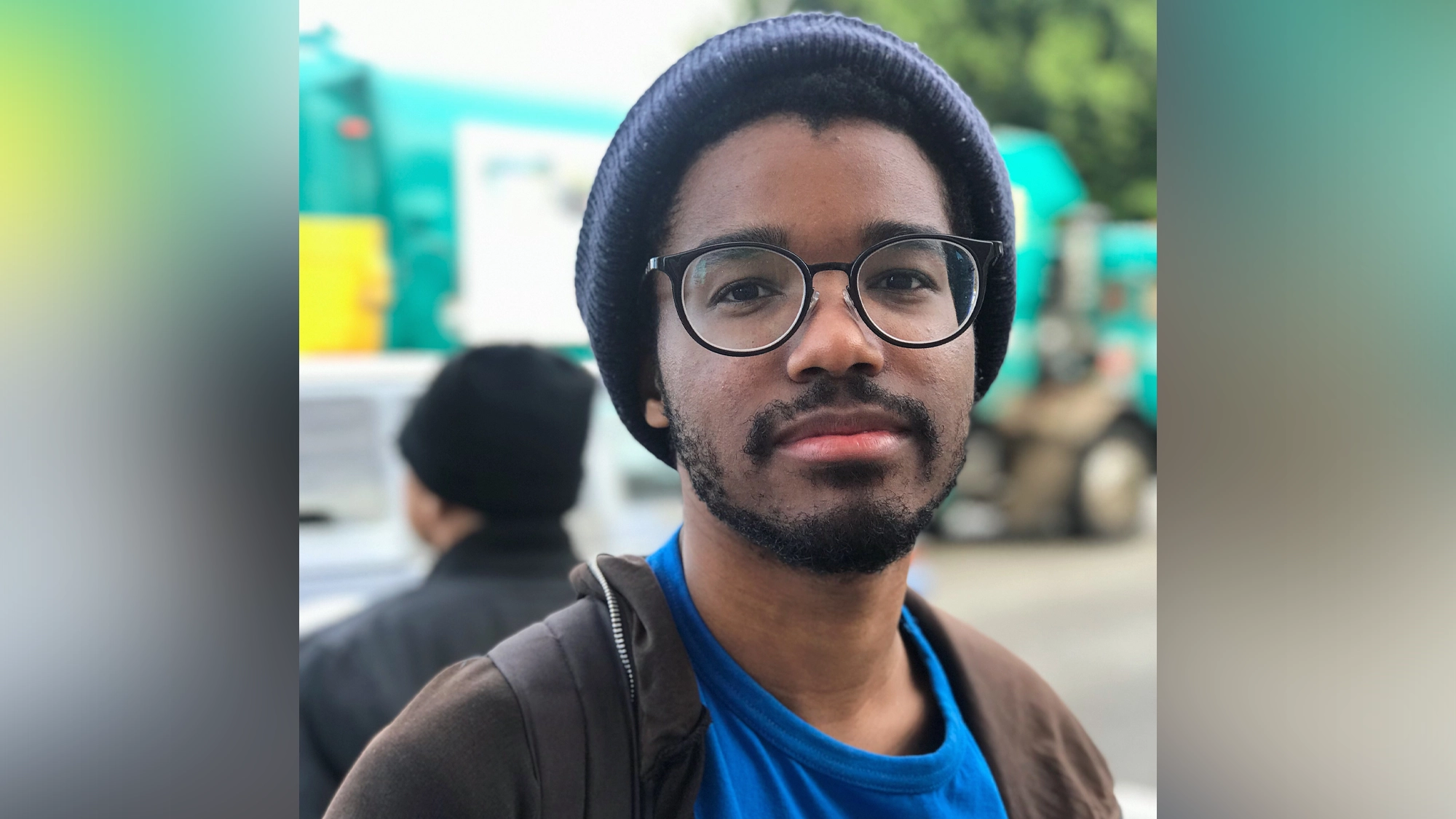 A young man with a short beard and mustache wears glasses, a dark beanie, a blue shirt, and a brown jacket. He looks into the camera with a neutral expression. The blurry background hints at an outdoor Khiyon In Concert event with teal vehicles.