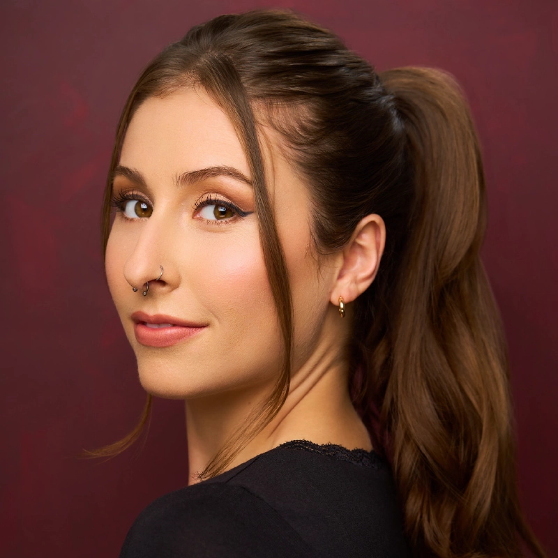 A young woman with long brown hair in a ponytail, wearing a black top. Kristin Weiss has winged eyeliner, natural makeup, and a septum piercing with a nose ring. She looks over her shoulder, smiling slightly, against a deep burgundy background.