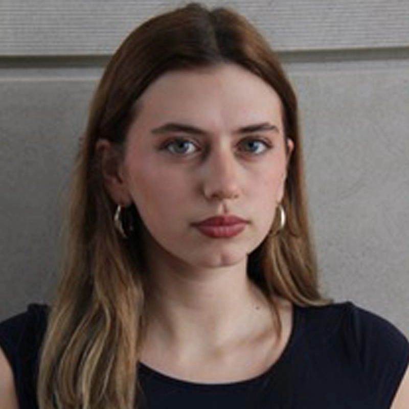 A young woman with fair skin and straight, light brown hair wears a black top and large hoop earrings. She looks directly at the camera with a neutral expression. The background has horizontal lines—photographed in the style of Allison Kuhn.
