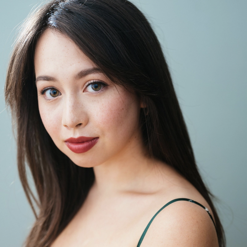 A headshot of Charlotte Kunesh, a woman with long, dark hair and light skin, looking directly at the camera with a gentle expression. She has hazel eyes, light freckles, and wears bold red lipstick. She is dressed in a dark green tank top against a simple, muted blue-grey background.