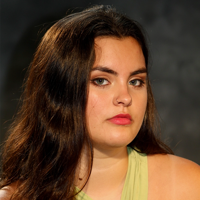 A young woman with long dark brown hair and light skin gazes seriously at the camera. Maya Monteleone wears a light green sleeveless top and a nose ring. The dark, blurred background draws attention to her neutral expression and face.