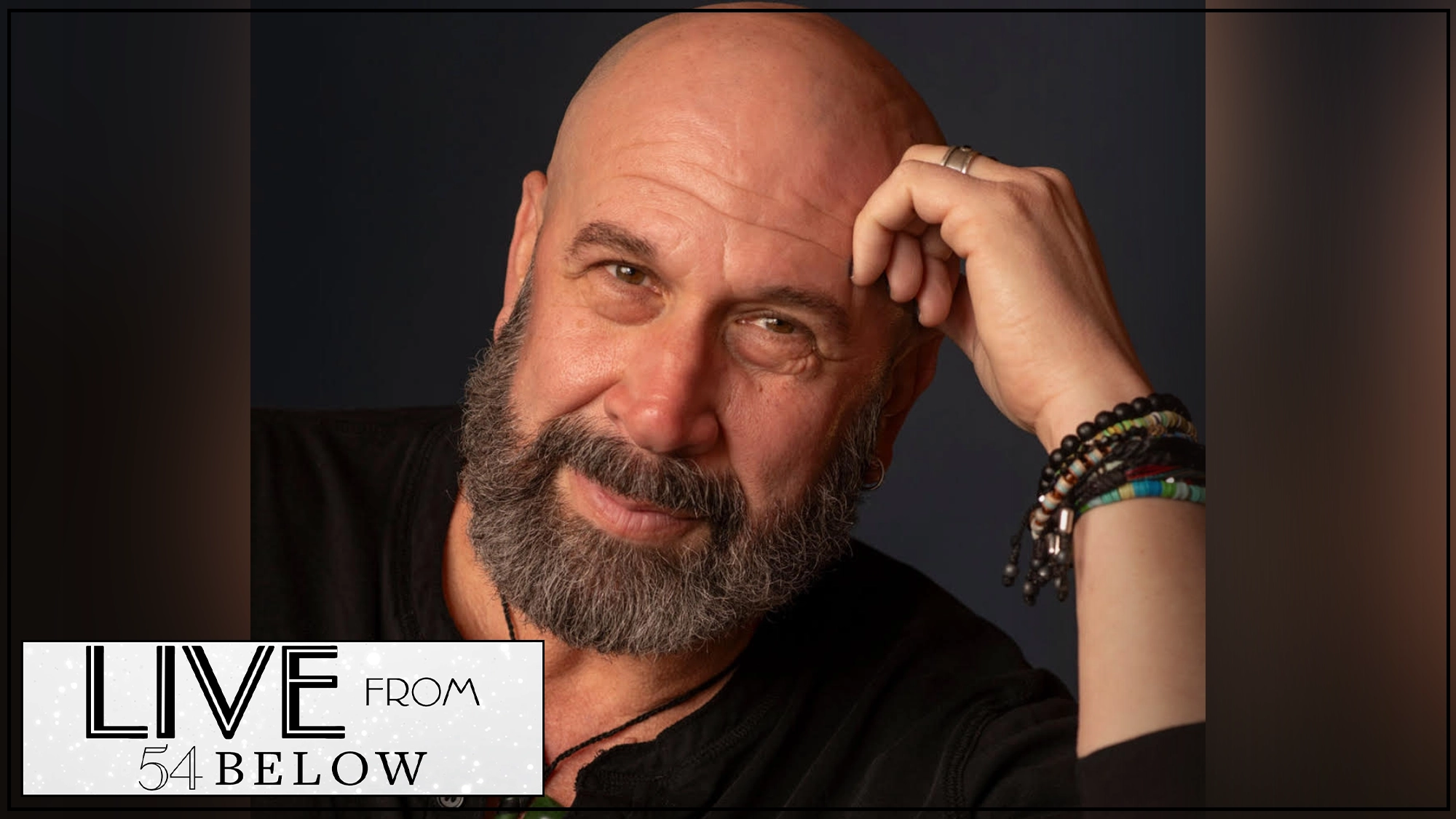 A bald, bearded man wearing a black shirt and colorful beaded bracelets leans his head on his hand, gazing at the camera with a gentle smile. In the bottom left, 