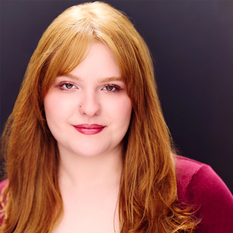 A young woman with long, straight red hair and fair skin smiles gently. She has a nose piercing and wears natural makeup with berry lipstick. She is dressed in a burgundy top and is posed against a plain dark background.