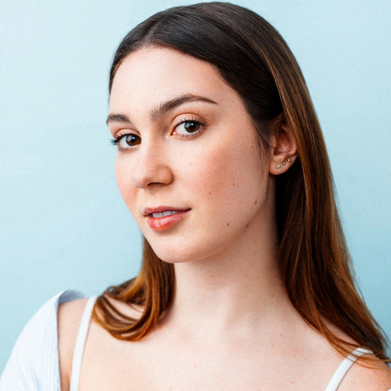 Zoey Mae-Dillon Levine, a young woman with long, straight brown hair and light skin, looks at the camera with a subtle smile. She wears a light-colored top and has multiple earrings in her ear against a solid pale blue background.