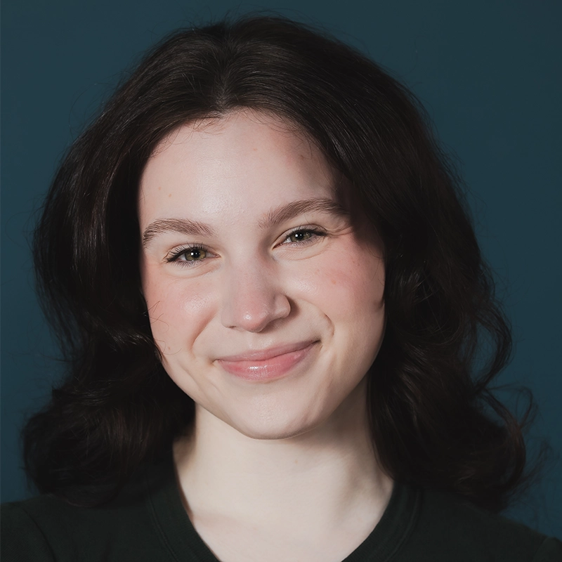 Gina Marie Incandela, a young woman with fair skin and wavy dark brown hair, smiles softly. She wears a dark top against a plain, dark blue background. Her minimal makeup and warm, friendly expression add to her approachable appearance.