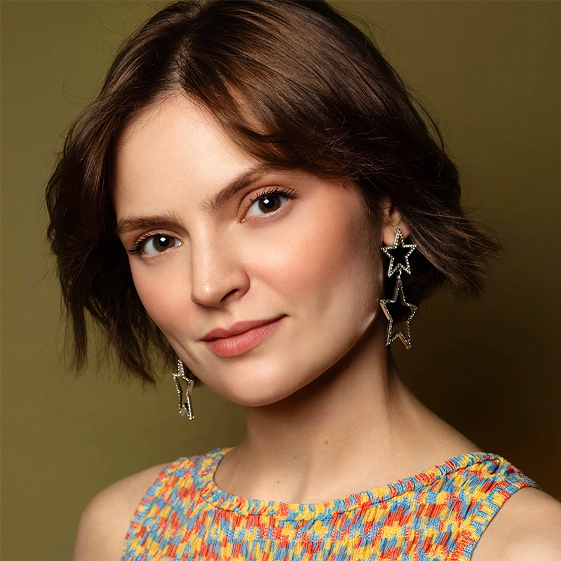 Jenna Leigh Miller, with short brown hair, wears a sleeveless multicolored top and large dangling star-shaped earrings. She smiles softly at the camera, posed against an olive green background.