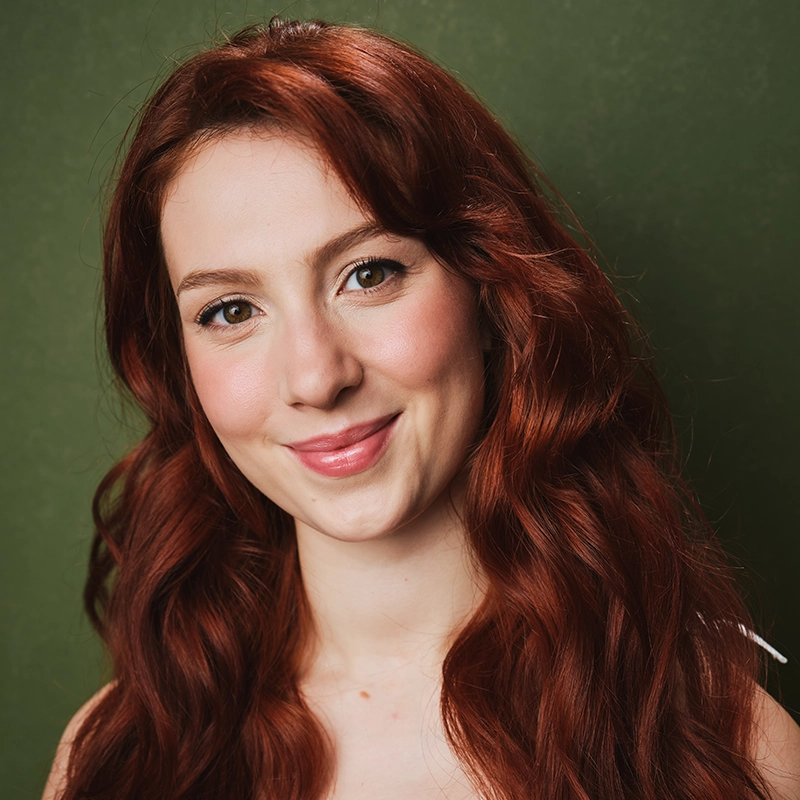 A young woman with long, wavy red hair and fair skin smiles softly at the camera. With light makeup, Mireya Velasquez is posed in front of a plain olive-green background, natural lighting accentuating her features and vibrant hair color.