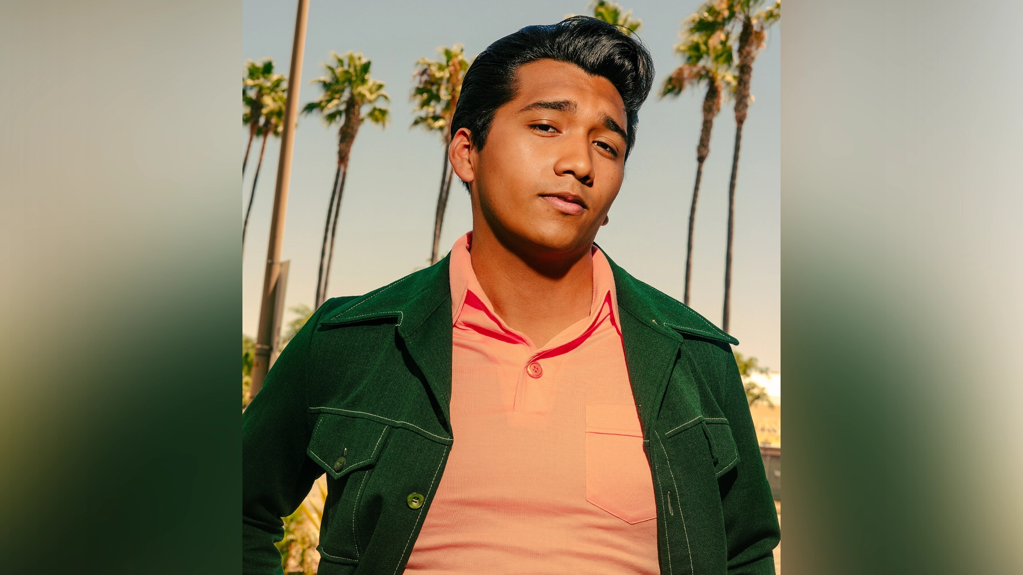 A young man with dark slicked-back hair wears a green jacket over a peach shirt. He stands outdoors in front of tall palm trees and a clear sky, looking confidently at the camera—ready to explore fabulous places under the warm, sunny ambiance.