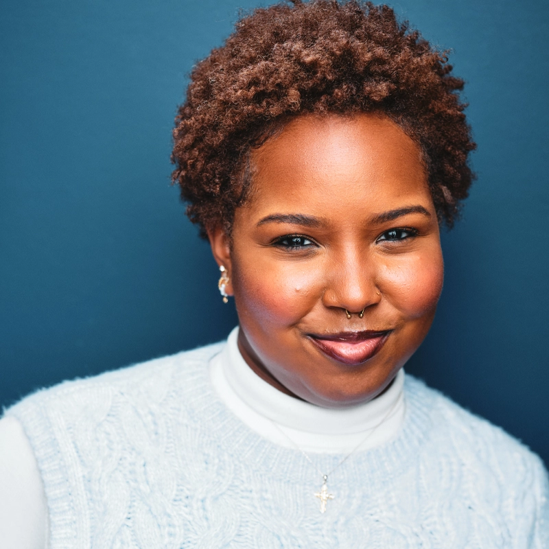 Judah Nicole Lyles, a woman with short, curly hair, smiles softly. She wears a light blue cable-knit sweater over a white turtleneck, a small gold cross necklace, earrings, and a septum ring. The background is a solid dark blue.