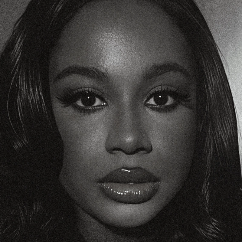 Black and white close-up of a woman with long, wavy hair. She has large eyes with dramatic lashes, defined brows, and full lips with glossy lipstick. The lighting highlights her smooth skin and symmetrical features, giving a classic, elegant look.