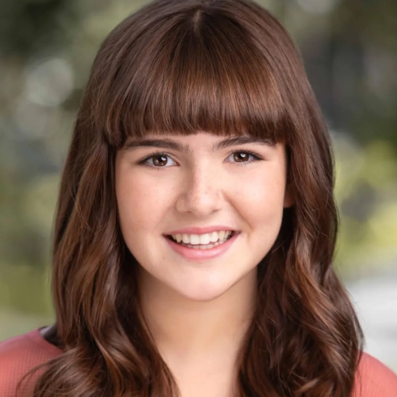 A young woman with long brown hair and bangs smiles at the camera. Jordan Gulliford wears a mauve top, and the softly blurred greenery in the background gives this portrait a warm, natural feel.