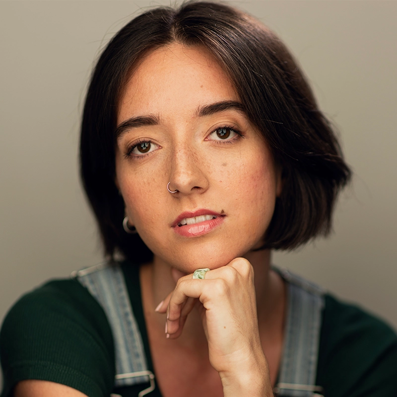 A young woman with short dark hair and a nose ring gazes softly at the camera. She rests her chin on her hand, wearing a green top and denim overalls. The background is neutral and softly blurred, highlighting her calm, thoughtful expression.