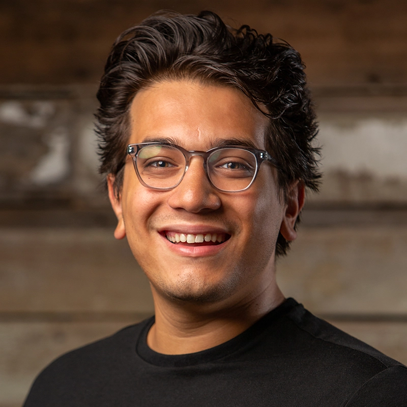 A young man with dark, wavy hair and glasses smiles warmly at the camera. He is wearing a black crew-neck shirt and stands in front of a blurred, wooden background, creating a friendly and approachable atmosphere.