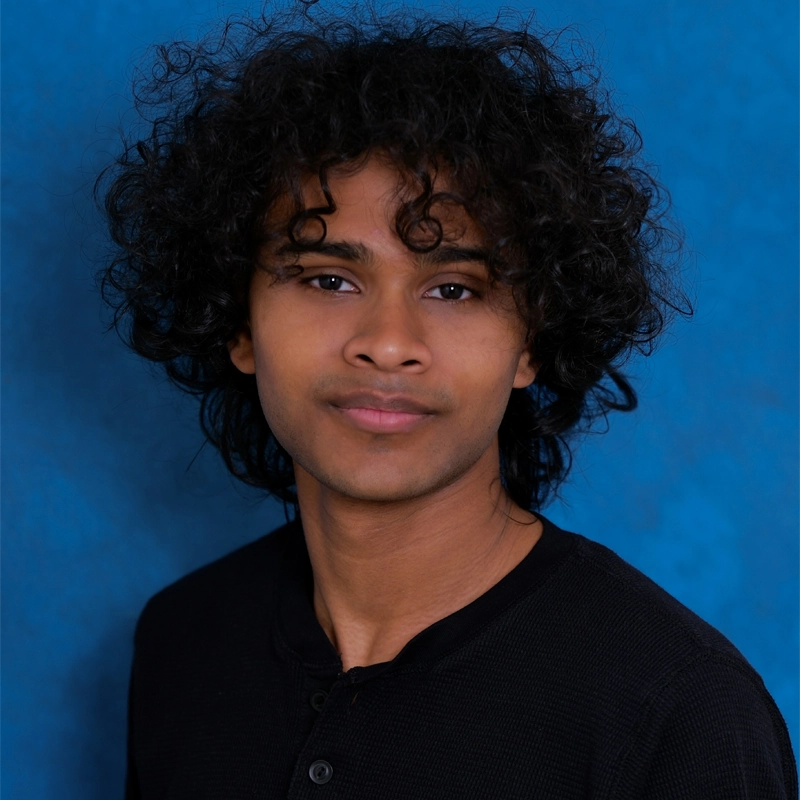 A young person with medium brown skin and thick, curly dark hair poses against a textured blue background. Wearing a black, buttoned shirt, Rakesh Mangru looks confidently at the camera with a gentle, neutral expression.