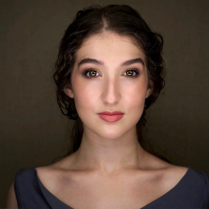 A young woman with wavy brown hair and fair skin, Milena Manocchia, looks directly at the camera. She wears natural makeup with pink lipstick and soft purple eyeshadow, her calm expression framed by a plain, dark background and a gray top.