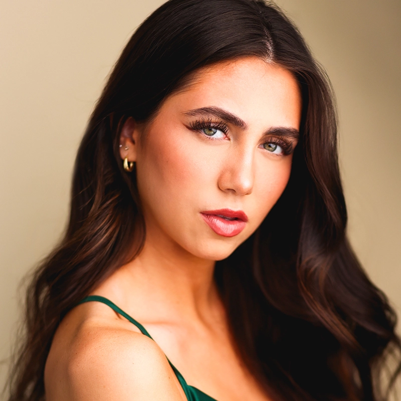 Maria Uicker, a young woman with long, wavy dark brown hair and green eyes, looks confidently at the camera. She wears subtle makeup, gold hoop earrings, and a green sleeveless top against a softly lit, professional backdrop.
