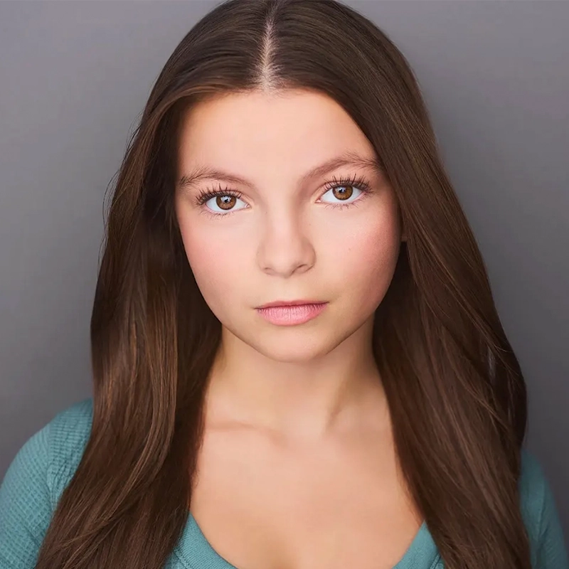 A young woman with straight, long brown hair and fair skin looks directly at the camera with a neutral expression. She wears a teal top, reminiscent of styles seen in Jake Hamilton’s portraits, standing before a plain grey background.