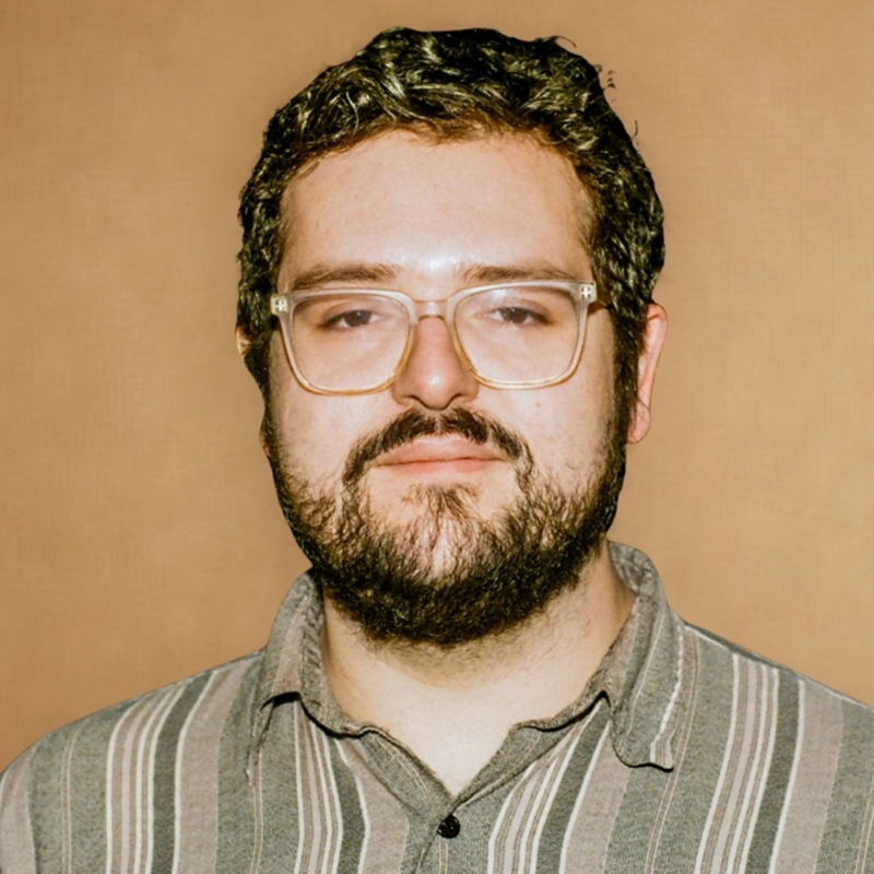 A man with short, wavy dark hair, a full beard, and mustache stands against a plain tan background. Wearing large clear-framed glasses and a gray striped collared shirt, Mario Fuentes looks directly at the camera with a neutral expression.