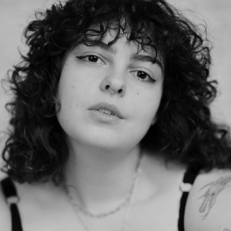 Black and white close-up of a person with curly dark hair, a nose ring, and winged eyeliner. They wear a thin-strapped top and a necklace. Their expression is neutral, and a tattoo is visible on their upper arm. The background is softly blurred.