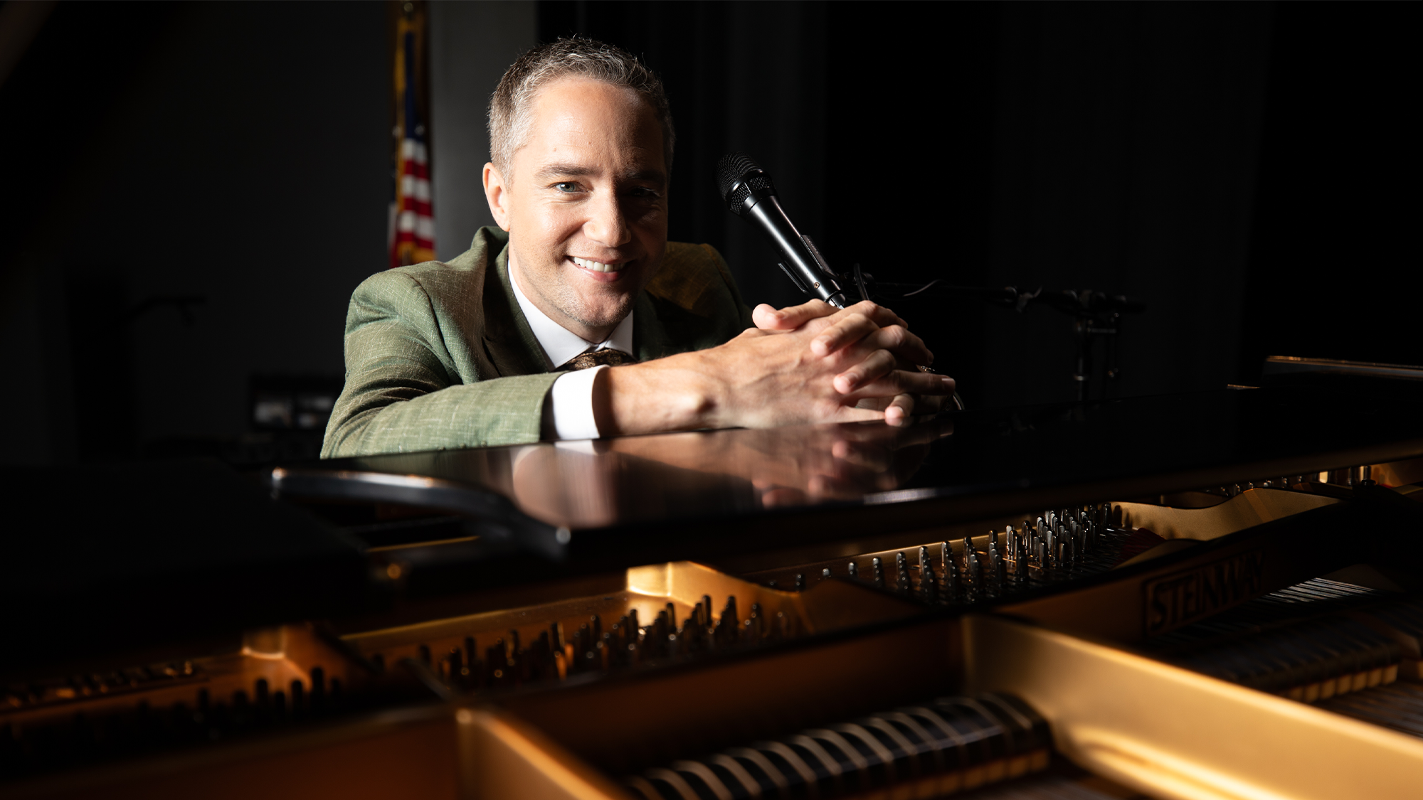 Marquis Studios A smiling man in a green suit sits at a grand piano with an open lid, holding a microphone close to his mouth. An American flag is visible in the background as warm light highlights the piano’s golden interior at the Marquis Studios Spring Benefit 2026.
