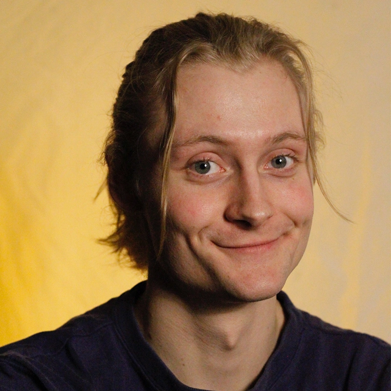 Chris Allyah, a young person with light skin and long blond hair tied back, smiles gently at the camera. They have blue eyes, visible dimples, and wear a dark shirt. The background glows softly with warm yellow tones.