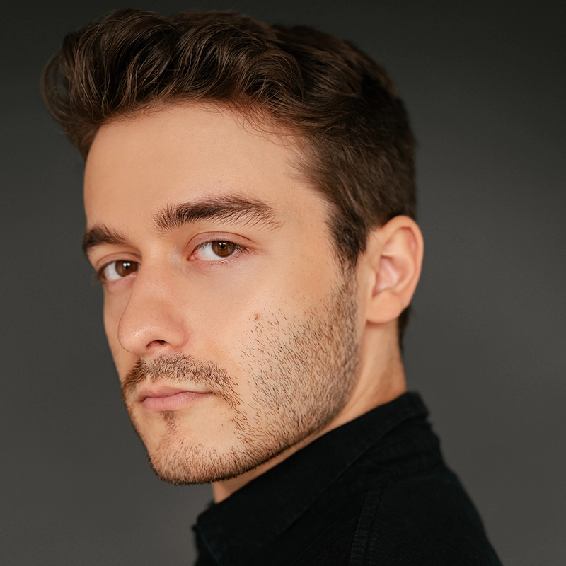 A young man with short brown hair and light facial stubble looks at the camera with a neutral expression. He wears a black shirt, posed against a simple dark gray background, in the soft, even lighting favored by Rachel Sklar’s portrait style.