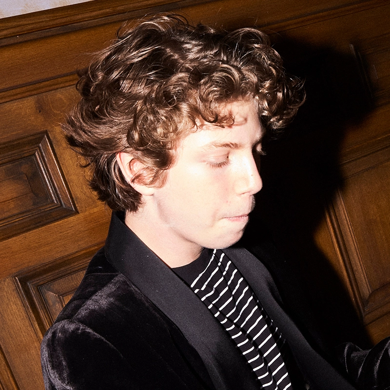 Matthew Brand, a young person with curly brown hair and fair skin, sits in profile against a wooden paneled wall. Eyes closed and lips pressed together, he wears a black velvet jacket over a black-and-white striped shirt, bathed in strong light.