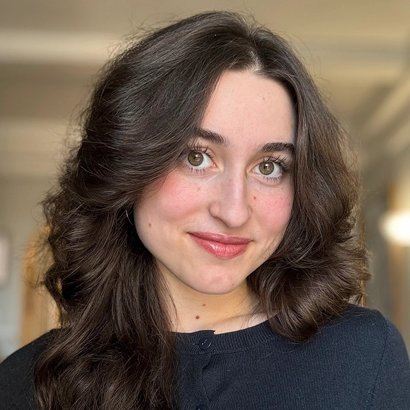 A young woman, Maya Monteleone, with long, wavy brown hair and green eyes smiles softly at the camera. She wears a black top and subtle makeup, with rosy cheeks and glossy lips. The background is softly blurred in natural light.