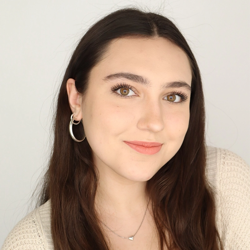 A young woman with long brown hair and hazel eyes smiles at the camera. She wears silver hoop earrings, a subtle necklace with a small pendant, a cream-colored knit top, and natural makeup. The background is plain and white.