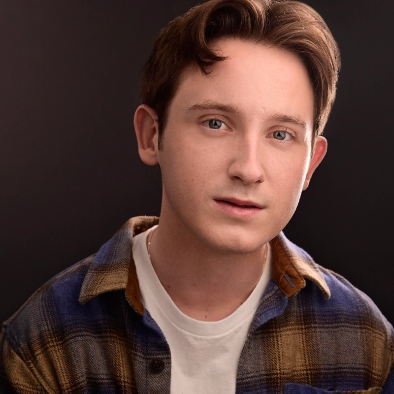 A young man with fair skin, short brown hair, and blue eyes looks at the camera. He wears a white t-shirt under a blue and brown plaid shirt. The background is dark and plain, highlighting his face and expression—introducing Luke McIntyre.