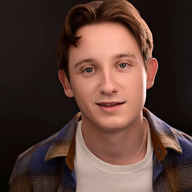 A young adult, Luke McIntyre, with fair skin, short light brown hair, and hazel eyes smiles gently. He wears a white t-shirt under a blue and brown plaid shirt against a dark, softly blurred background. Warm lighting highlights his face.