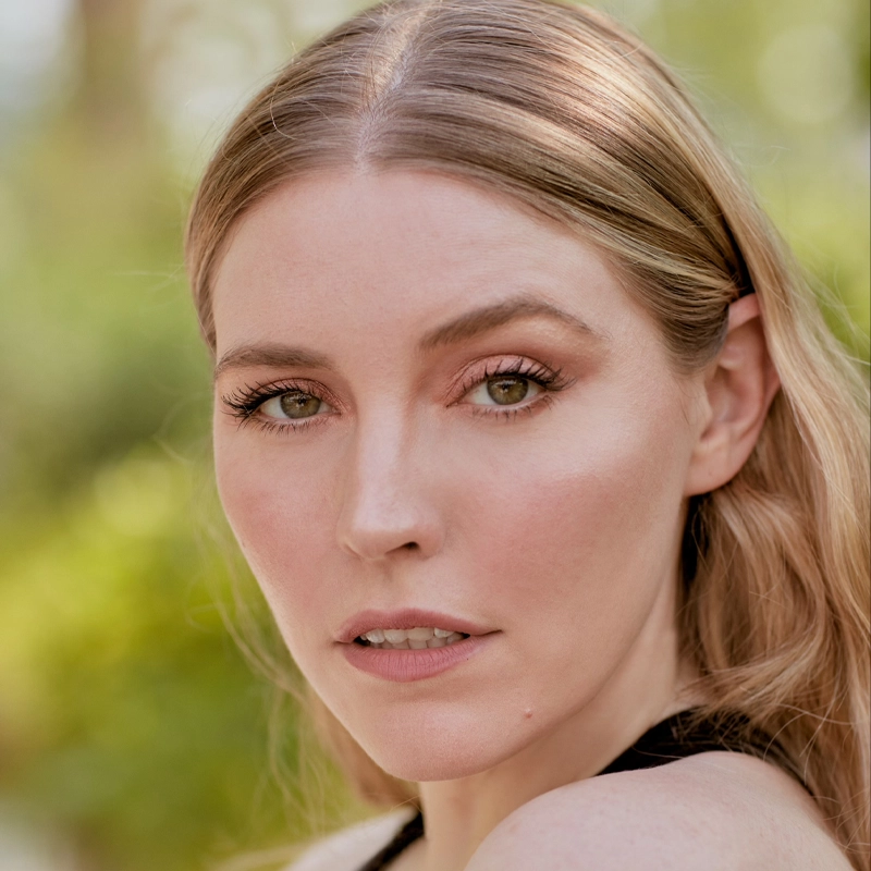 A woman with fair skin and long, wavy blonde hair looks directly at the camera. Cydney McQuillan-Grace has light hazel eyes, natural makeup, and slightly parted lips. The blurred greenery in the background gives a soft, outdoor feel to the portrait.