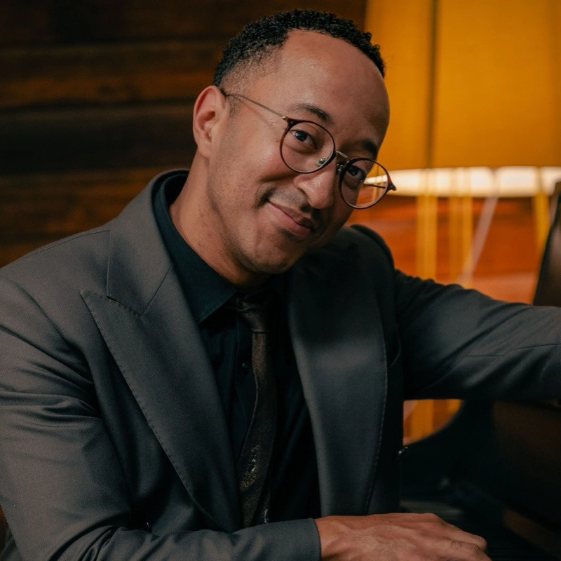 A man resembling Mark Meadows, with short curly hair, glasses, and a trimmed goatee, sits indoors in a gray suit, dark shirt, and patterned tie. He smiles warmly at the camera with one arm resting on a piano under soft yellow lamp light.