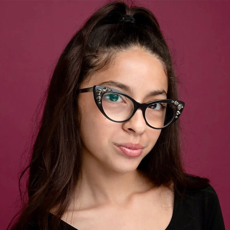 A young woman with long dark hair in a high ponytail wears black cat-eye glasses with silver studs. She has light skin, subtle makeup, and a black top, posed against a solid maroon background—an image styled by Jake Hamilton.