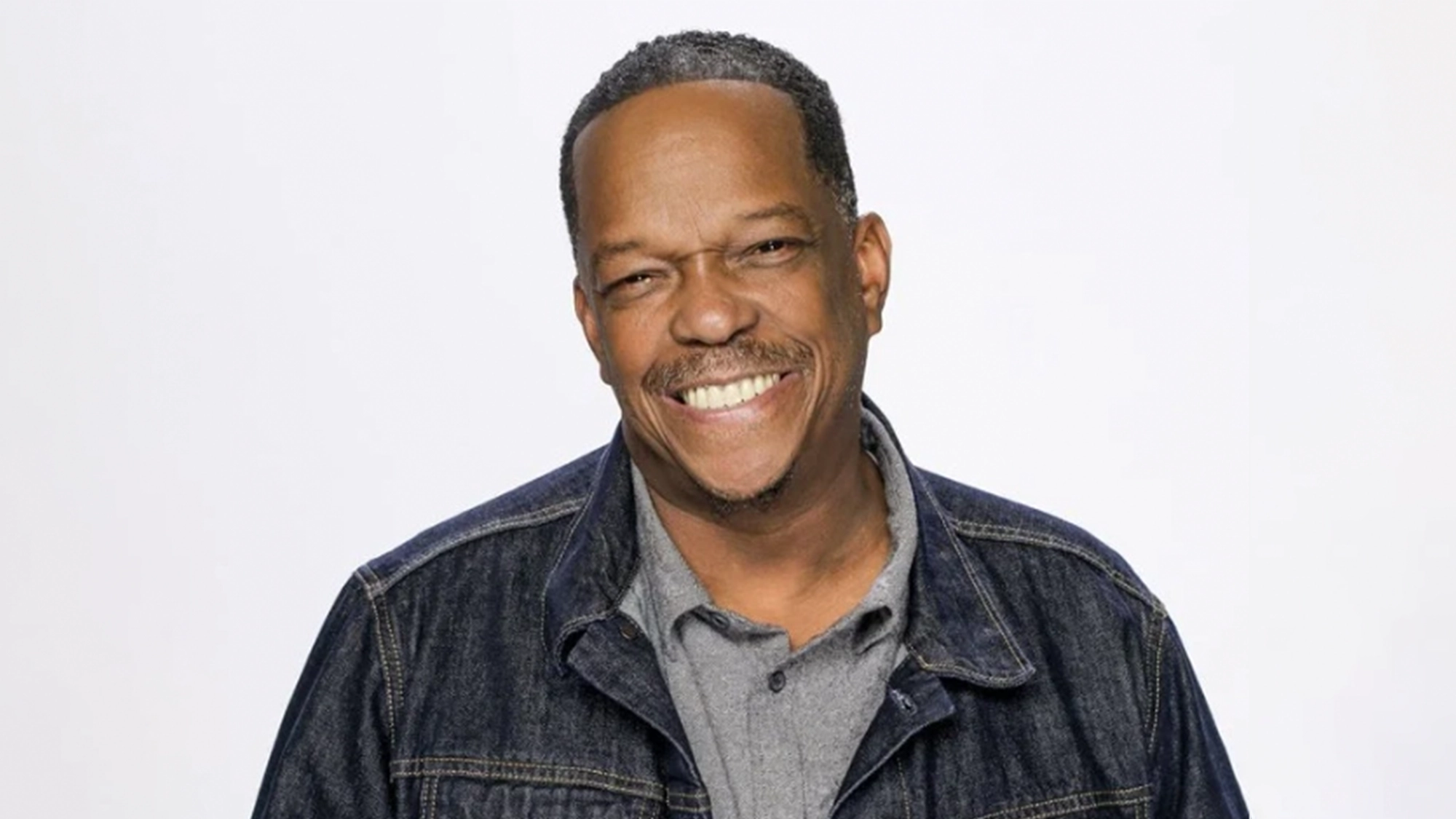 A middle-aged man with short hair and a mustache smiles warmly at the camera. He is wearing a dark denim jacket over a gray collared shirt, standing against a plain white background.