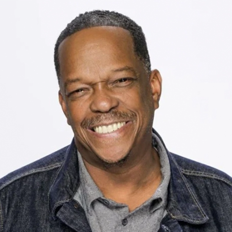 A middle-aged Black man with short hair and a mustache smiles warmly at the camera. He wears a gray polo shirt under a blue denim jacket. The background is plain and light-colored, drawing focus to his cheerful expression.