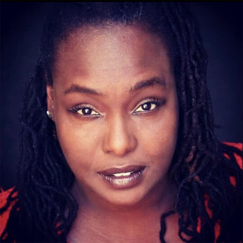 Dee-D Miller, with dark skin and long, twisted hair, looks directly at the camera with a neutral expression. Wearing a small earring and red top against a black background, their wide eyes and slightly parted lips draw you in.