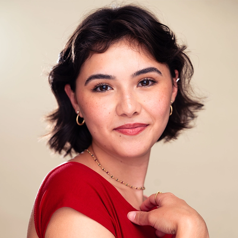 A young woman with short dark wavy hair smiles gently, wearing a red top, gold hoop earrings, and a beaded necklace. Mireya Velasquez poses in front of a neutral beige background, with a soft expression and natural makeup.