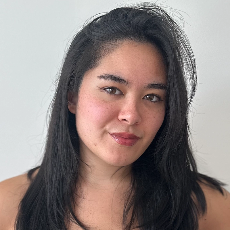 A woman with long black hair and fair skin gazes at the camera, wearing subtle makeup with winged eyeliner and natural pink lipstick. She stands against a plain white background and smiles softly, creating a friendly and approachable expression.