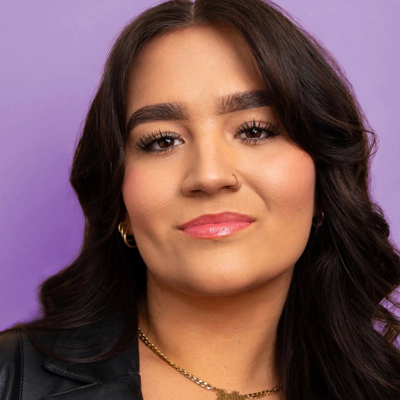 A young woman with long, dark wavy hair poses against a purple background. She wears a black jacket, gold hoop earrings, a gold necklace, a nose ring, and glossy pink lipstick. Her expression is confident and relaxed.