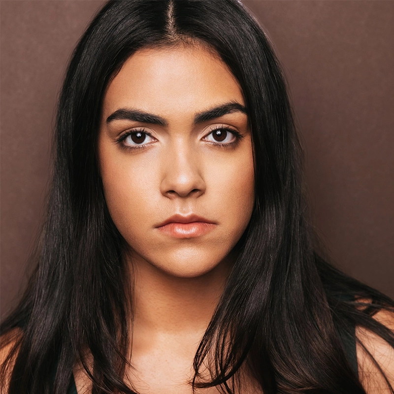 Maya Monteleone, a young woman with long, straight dark hair and thick eyebrows, looks directly at the camera with a neutral expression. She has smooth, medium-toned skin, natural makeup, and is set against a plain brown background.