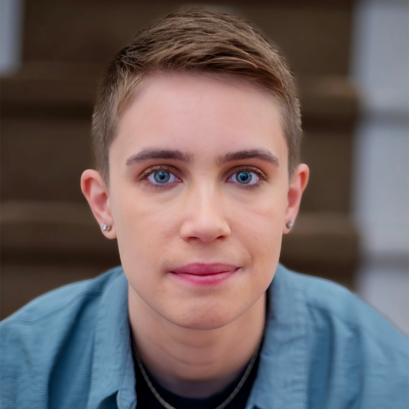 A person with short light brown hair and blue eyes looks directly at the camera. They are wearing a blue shirt, small earrings, and a necklace. The background is softly blurred steps in shades of brown and gray.