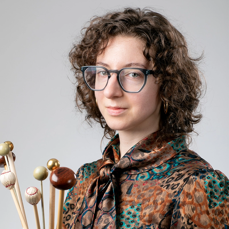 A young person with curly brown hair and glasses wears a colorful, patterned blouse with a bow tie. Mario Fuentes holds a group of mallets with different colored heads. The background is plain light gray, and the expression is calm and confident.