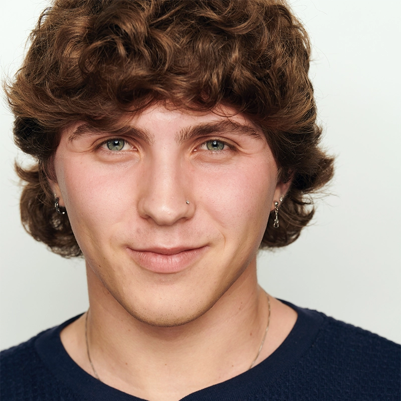 A young person with wavy brown hair, light skin, and green eyes smiles softly at the camera. They wear small hoop earrings, a nose ring, a thin chain necklace, and a navy textured shirt. The background is plain white.