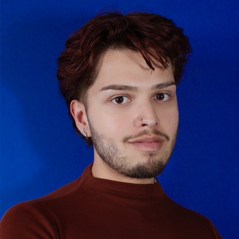 Maria Uicker, a young person with short, wavy brown hair, a beard, and a nose ring, poses against a solid blue background. They wear a brown high-neck top and have a neutral expression, looking directly at the camera.