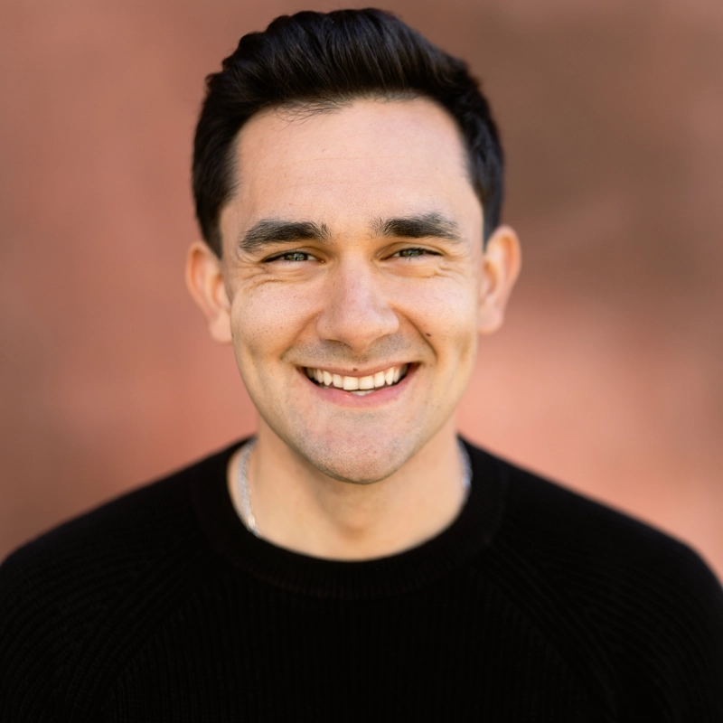 A man with short dark hair and light skin smiles at the camera. He is wearing a black sweater. The softly blurred warm brown background keeps Shane Nourie as the clear focus of the image.
