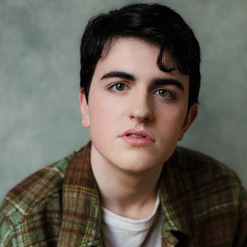 A young person, Finn O’Rourke, with short dark hair and hazel eyes gazes softly at the camera. They wear a green and brown plaid shirt over a white top, set against a muted, mottled gray-green background that creates a calm and neutral backdrop.