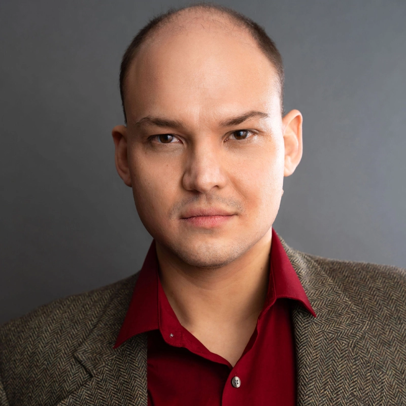 Michael Pacholski, a man with light skin, closely shaved dark hair, and a receding hairline, looks directly at the camera with a neutral expression. He wears a red dress shirt and a brown textured blazer against a plain gray background.