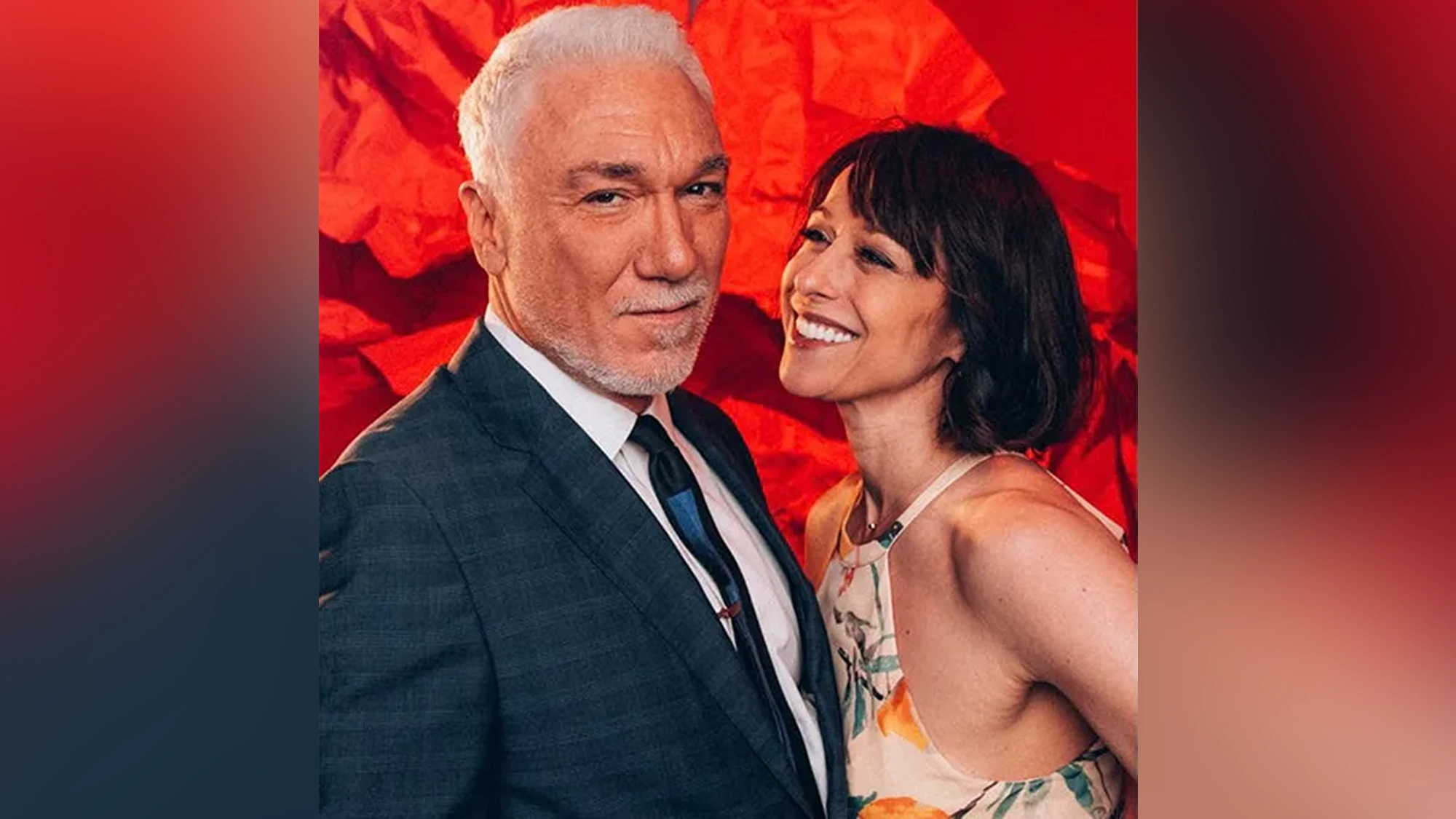Patrick Page, in a dark checkered suit and tie, stands beside Paige Davis, who wears a sleeveless floral dress. Together, they pose against a bright red backdrop—happy and relaxed—a love story captured in a single moment.
