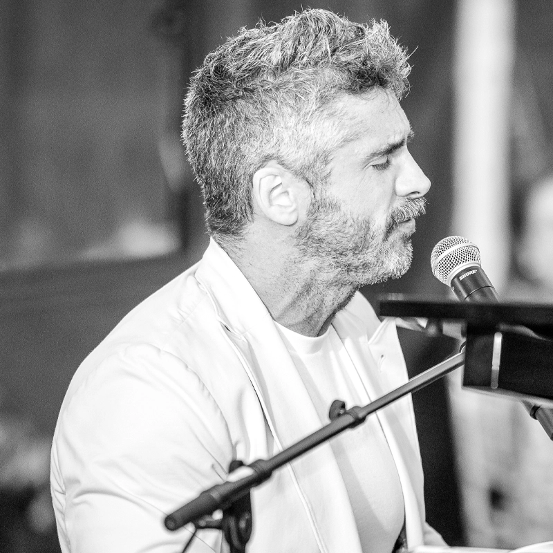 A grayscale photo of a man with short, wavy gray hair and a beard, wearing a light jacket and T-shirt, singing into a microphone while playing piano. He appears focused, with eyes closed, in a close-up side profile view.